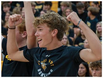A person with curly hair wearing a black shirt is cheering with raised arms in a crowded indoor setting. Descripci n generada con IA