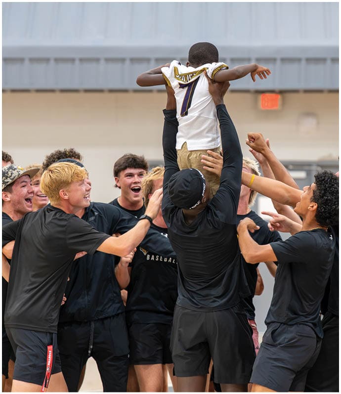 A group of people wearing black athletic clothing are joyfully lifting one person into the air inside a gymnasium. Descripci n generada con IA