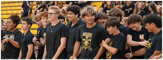 A group of people wearing black shirts with a yellow design are standing closely together in a gymnasium with yellow bleachers in the background. Descripci n generada con IA