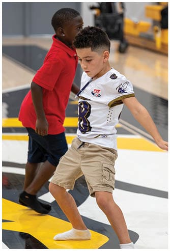 A child wearing a sports jersey and shorts is actively playing on an indoor court with another child in a red shirt nearby. Descripci n generada con IA
