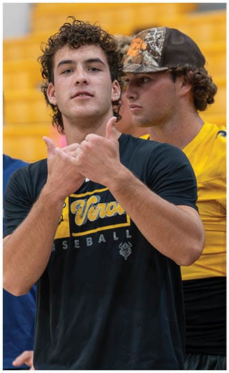 Two adults stand in a sports setting with one wearing a black baseball shirt making a hand gesture and the other wearing a yellow sports jersey and a camouflage cap looking to the side. Descripci n generada con IA