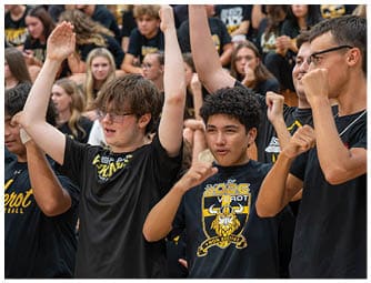 A group of people wearing black shirts with yellow designs are raising their arms and cheering enthusiastically in a crowded setting. Descripci n generada con IA