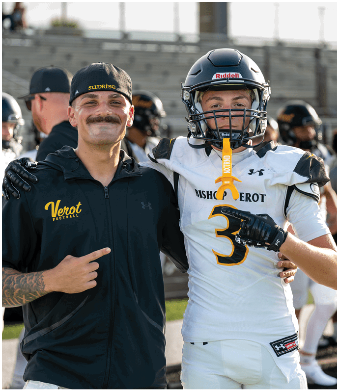 Two people stand on a sports field with one wearing a football uniform and helmet holding a mouthguard while the other wears a black jacket and cap, both smiling and posing for the photo. Descripci n generada con IA