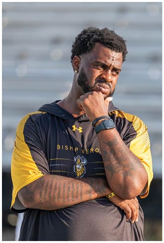 A person wearing a black and yellow Bishop Verot sports jersey stands with arms crossed and a thoughtful expression while resting their chin on one hand. Descripci n generada con IA