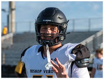 A person wearing a black football helmet and a white and black football jersey is smiling on a sports field with empty bleachers in the background. Descripci n generada con IA