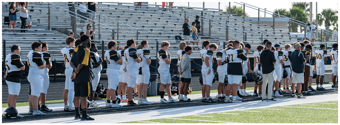 A group of people wearing sports uniforms and casual clothing stand in a line on the edge of a sports field. Descripci n generada con IA