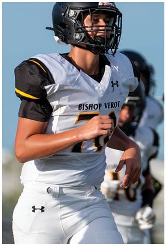 A person wearing a black helmet and a white football uniform with “Bishop Verot" on the jersey is running on a field with another player blurred in the background. Descripci n generada con IA