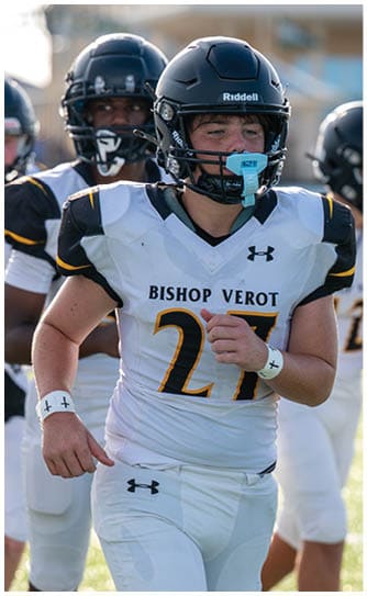 A group of people in football uniforms and helmets are running on a field with one person in the foreground wearing a white jersey with “Bishop Verot" and the number 27. Descripci n generada con IA