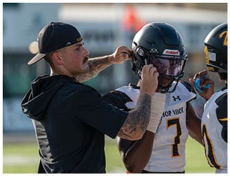 An adult wearing a backward cap adjusts the helmet of a person in a football uniform with the number seven on it during a practice or game. Descripci n generada con IA