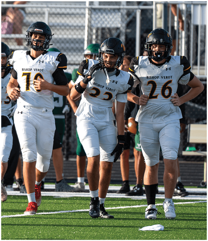 Three people wearing white football uniforms with helmets walk together on a green field during a game or practice. Descripci n generada con IA