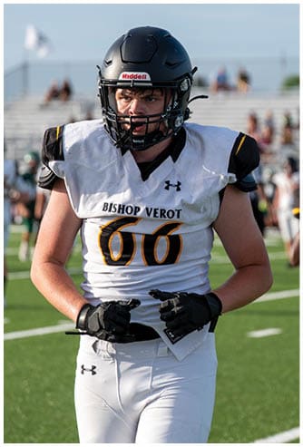 A person wearing a black football helmet and white Bishop Verot jersey with the number 66 stands on a football field with blurred players and bleachers in the background. Descripci n generada con IA