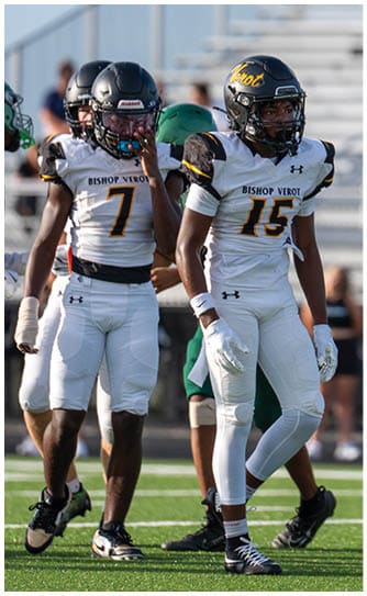 Two people in white and black football uniforms with helmets stand on a green field while other players and spectators are visible in the background. Descripci n generada con IA
