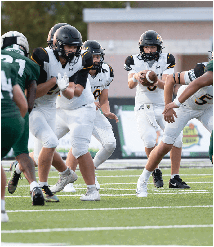 People in sports uniforms are actively playing football on a field with yard lines visible. Descripci n generada con IA