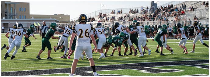 People in sports uniforms are playing football on a field with spectators seated in bleachers in the background. Descripci n generada con IA
