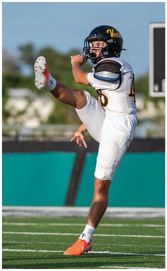 A person wearing a football uniform is kicking a ball on a grassy field with one leg raised high and the other planted on the ground. Descripci n generada con IA