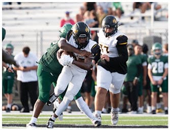 Two people in football uniforms are engaged in a tackle on a field while another player in uniform runs nearby with spectators in the background. Descripci n generada con IA