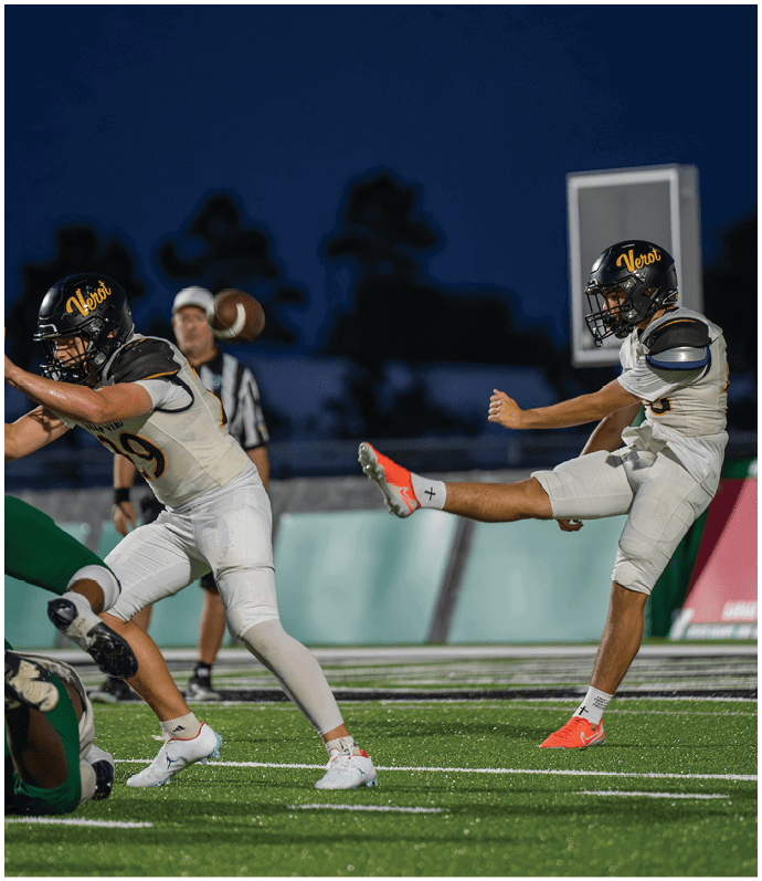 Two people in football uniforms are actively playing on a field at night, with one kicking the ball and the other preparing to block. Descripci n generada con IA