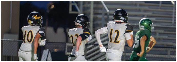 Four people in football uniforms are running on a field with one person in a green uniform holding a football and three others in white uniforms chasing behind. Descripci n generada con IA