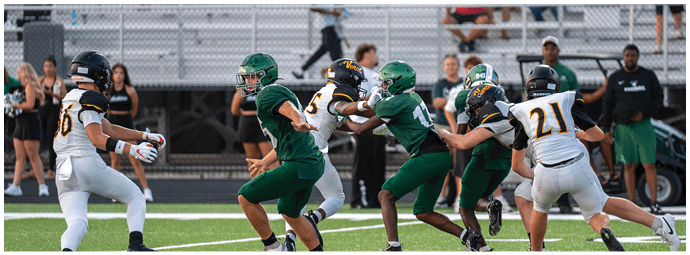 People in football uniforms are actively engaged in a play on a field with bleachers and spectators in the background. Descripci n generada con IA