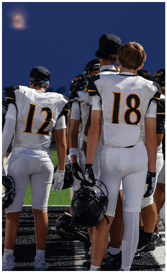 People wearing white football uniforms with numbers 12 and 18 stand on a field holding helmets while facing away from the camera. Descripci n generada con IA