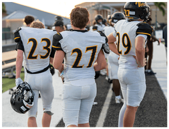 Three people wearing white football uniforms with black and yellow accents stand on the sidelines of a field, facing away from the camera. Descripci n generada con IA