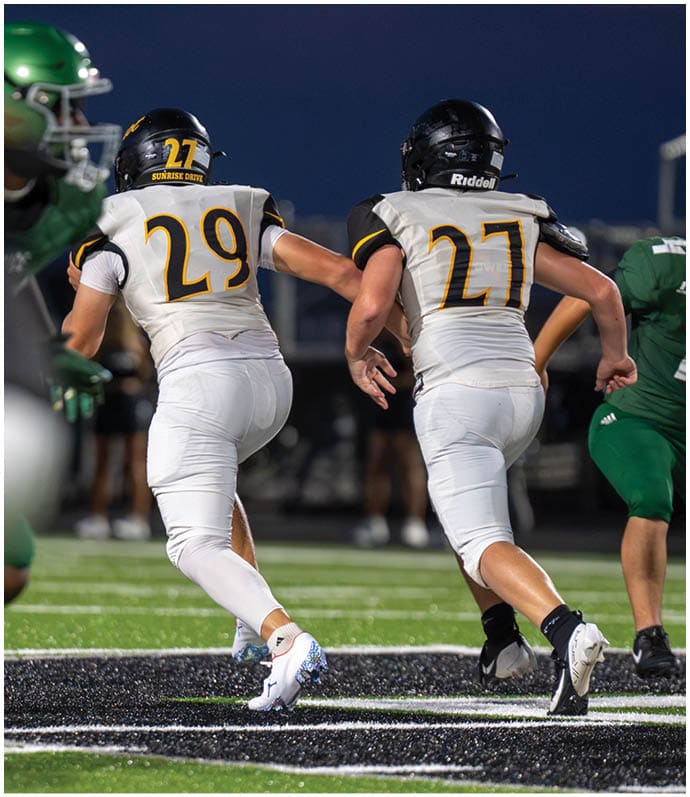 Two people in white sports uniforms with black helmets are running on a football field while two others in green uniforms are nearby. Descripci n generada con IA