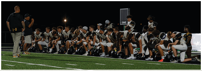 A group of people in football uniforms are kneeling on a field at night while two people stand nearby, one speaking into a microphone. Descripci n generada con IA