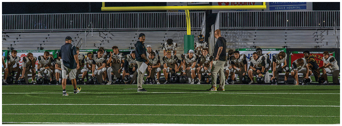 A group of people in sports uniforms and casual clothing are gathered on the sidelines of a football field, some standing and some sitting, with a goalpost and bleachers in the background. Descripci n generada con IA
