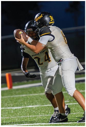 Two people in football uniforms are engaged in a play on a field, with one holding the ball and the other preparing to receive or block. Descripci n generada con IA