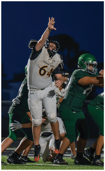 People in football uniforms are actively engaged in a nighttime game on the field with one player in white attempting to block or reach over players in green. Descripci n generada con IA