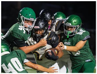 Several people wearing football helmets and uniforms are engaged in a tackle during a nighttime game on a field. Descripci n generada con IA