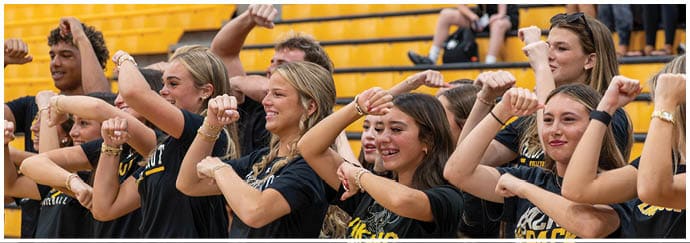 A group of adults wearing matching black shirts are standing in a row with their arms raised and fists clenched, appearing to cheer or chant in a stadium with yellow bleachers. Descripci n generada con IA