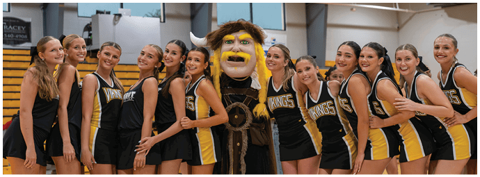 A group of people in matching black and yellow sports uniforms stand closely together smiling with a person in a large mascot costume in the center inside a gymnasium. Descripci n generada con IA
