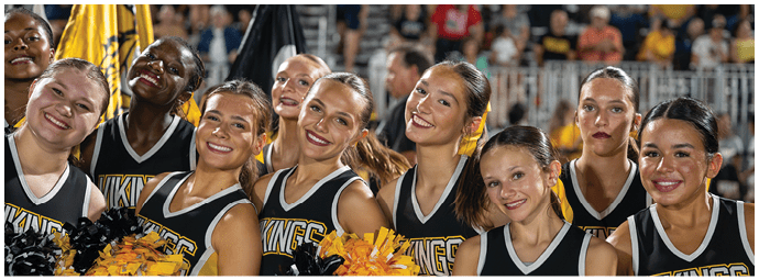 A group of smiling people wearing matching cheerleading uniforms with “VIKINGS" on the front are standing closely together holding yellow and black pom-poms at an outdoor event. Descripci n generada con IA