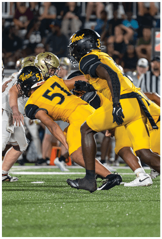 Three football players in yellow and black uniforms are crouched and ready to start a play on a green field with a crowd and referees in the background. Descripci n generada con IA