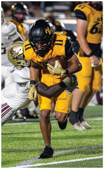 A person in yellow football gear is running with the ball while another person in white football gear dives to tackle them on a grassy field during a game. Descripci n generada con IA