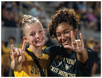 Two adults smiling and making hand signs while standing close together at an outdoor event with blurred spectators in the background. Descripci n generada con IA