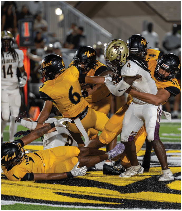 Several football players in yellow and white uniforms are engaged in a play on the field, with one player lying on the ground and others actively reaching or blocking. Descripci n generada con IA