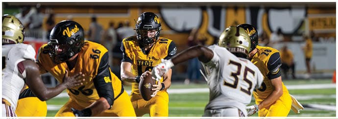 Adults in football uniforms are engaged in a game on a field with one player holding the ball and others preparing to block or tackle. Descripci n generada con IA