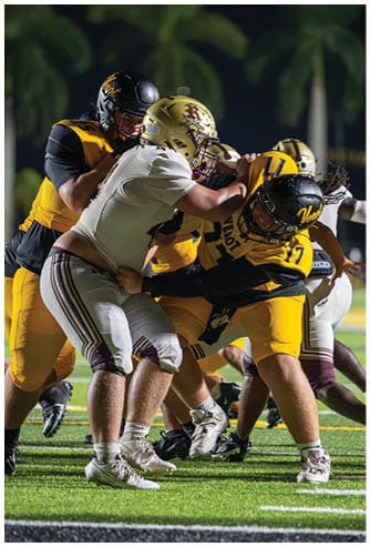 Several people wearing football uniforms are engaged in a close physical play on a grassy field under bright lights. Descripci n generada con IA