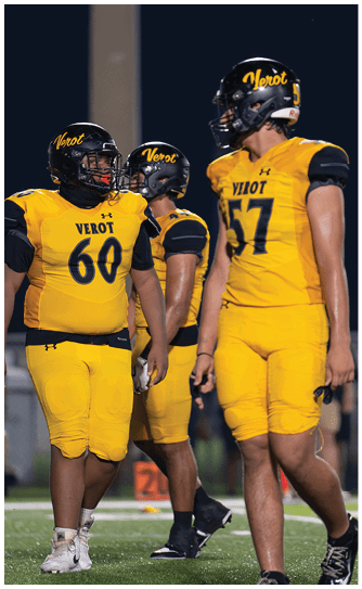 Three people wearing yellow and black football uniforms with helmets stand together on a field at night. Descripci n generada con IA