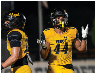 Two adults wearing yellow and black football uniforms with helmets are on a field at night, with one raising both hands as if communicating or reacting. Descripci n generada con IA