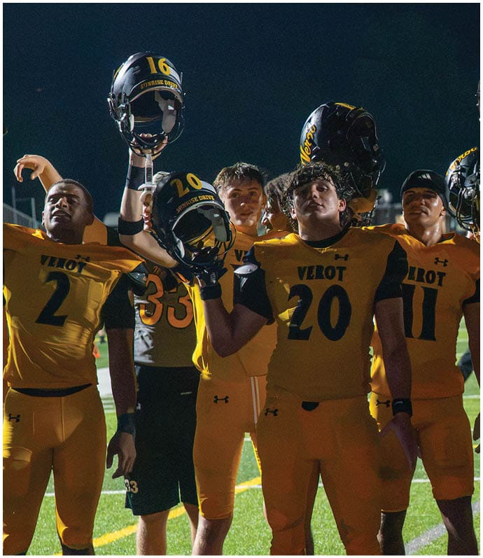 A group of people wearing yellow and black football uniforms stand together on a field at night, holding their helmets up in a celebratory gesture. Descripci n generada con IA