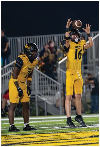 A person in a yellow football uniform is catching a football while other players in yellow and white uniforms watch on a field at night. Descripci n generada con IA