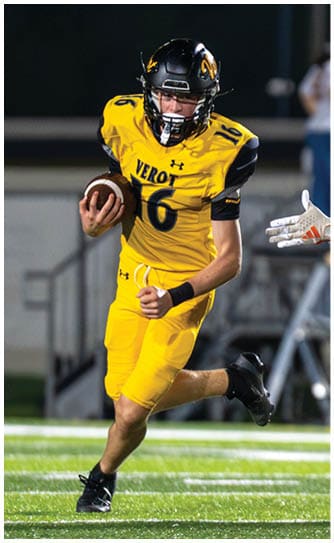 A person in a yellow football uniform is running with the ball while another person in a white football uniform is reaching out to try to tackle them on a lit football field. Descripci n generada con IA
