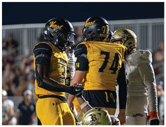Two football players in yellow and black uniforms stand on the field while a player in a white uniform kneels in front of them during a game with a crowd in the background. Descripci n generada con IA