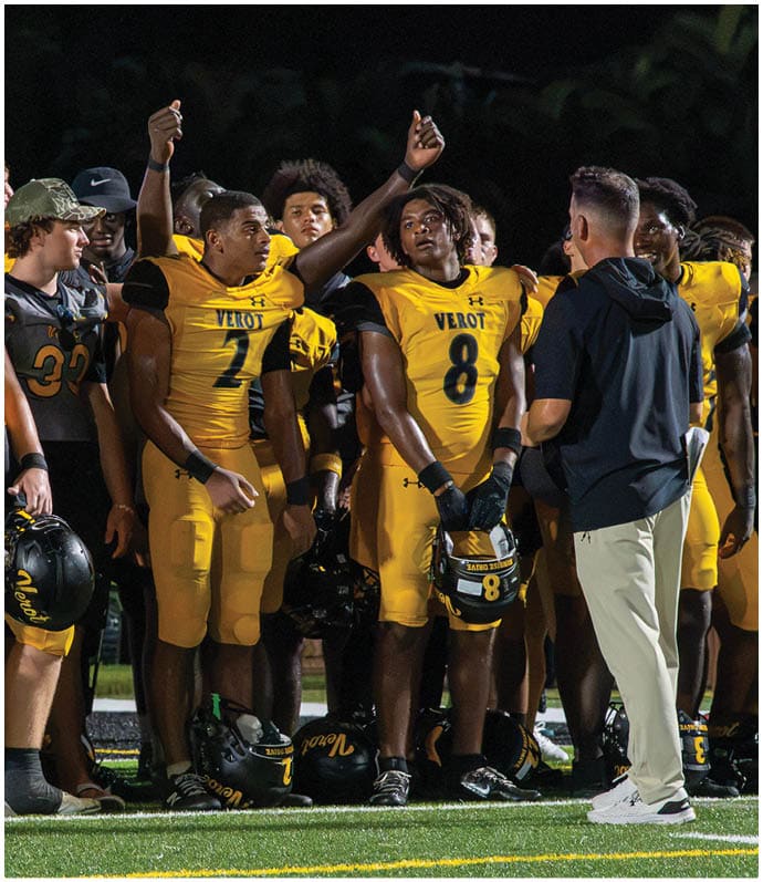 A group of people wearing yellow football uniforms stand together on a field at night, some holding helmets and others raising their hands. Descripci n generada con IA