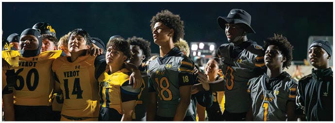 A group of people wearing football uniforms stand closely together on a field at night with stadium lights in the background. Descripci n generada con IA