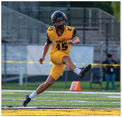 A person in a yellow and black football uniform is kicking a football on a field while another person in a white and maroon uniform watches. Descripci n generada con IA