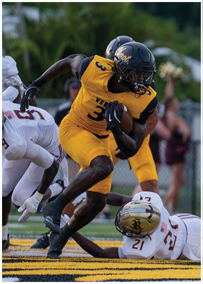 A person in a yellow sports uniform is running with a football while two people in white uniforms are attempting to tackle them on a football field. Descripci n generada con IA